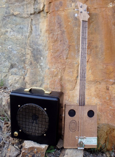 Flor De Tabaco wood cigar box guitar and black amplifier  from Wires and Wood resting up against a rock wall.