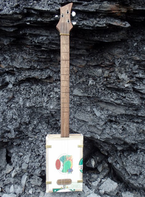 White Cuaba cigar box guitar from Wires and Wood in front of black rock wall.