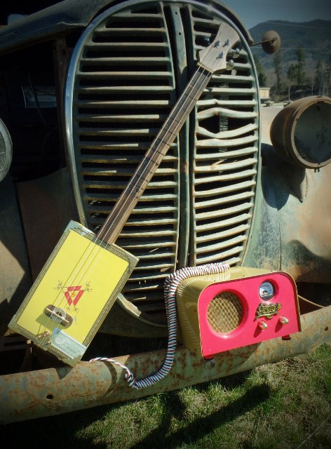 Monte Cristo Yellow cigar box guitar from Wires and Wood with a red amplifier on the front bumper of an old truck.