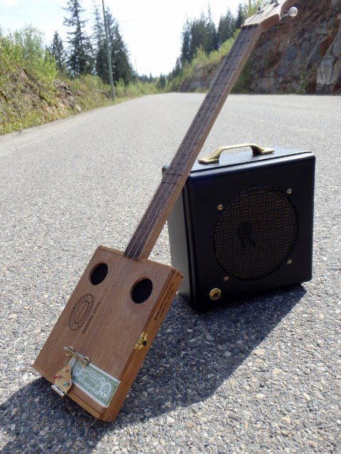 Wood Cigar Box Guitar from Wires and Wood resting on a black amplifier looking down a road.